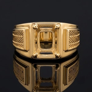 May include: A polished yellow gold ring setting featuring a rectangular center mount with four prongs designed to hold a gemstone. The band displays a textured herringbone pattern on both sides, providing a detailed finish to the metal structure.