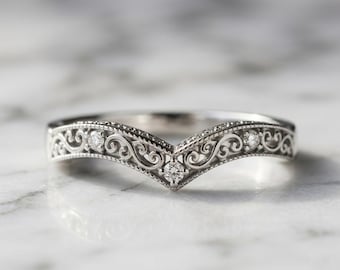 Puede incluir: Un anillo plateado en forma de chevrón con intrincados patrones de filigrana a lo largo de la banda. El anillo está acentuado con tres pequeñas piedras redondas, transparentes y facetadas en el centro y los lados del diseño en V.