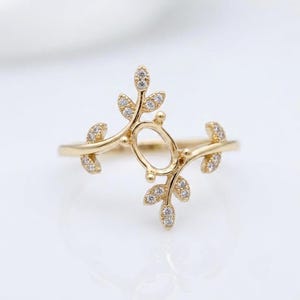 Puede incluir: Un anillo de oro con un engaste ovalado central enmarcado por dos ramas simétricas en forma de enredadera. Cada rama está adornada con pequeñas piedras claras y brillantes a lo largo de las hojas, creando un diseño delicado y natural.