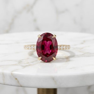 Puede incluir: Un anillo de oro con una gran piedra preciosa de color rojo intenso y corte ovalado, sujeta por cuatro garras. La banda es fina y está decorada con una hilera de pequeñas piedras claras de corte redondo engastadas en la mitad superior.