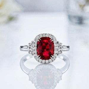 May include: A silver-toned ring featuring a large, oval-cut deep red gemstone at the center. The central stone is surrounded by a halo of small, clear, round-cut stones. Additional small clear stones are set along the band on both sides.