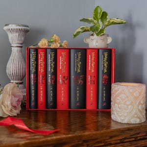 Puede incluir: Un set de siete libros de Harry Potter de J.K. Rowling en una caja roja. Los lomos alternan entre negro y rojo, con letras doradas e ilustraciones de leones. El conjunto está dispuesto horizontalmente sobre una superficie de madera.