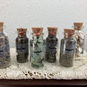 Peut inclure: Six flacons en verre transparent avec des bouchons en liège scellés à la cire cuivrée. Cinq flacons contiennent des herbes séchées étiquetées Lavender, Lemon Balm, Eucalyptus, Rosemary et Mugwort. Le sixième contient des petits coquillages blancs.