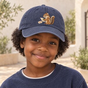 Peut inclure: Une casquette de baseball bleu marine ornée d'une broderie représentant un tamia brun tenant un crayon vert et écrivant sur une feuille de papier blanche, avec une petite icône d'enveloppe située à gauche du tamia sur le panneau avant.