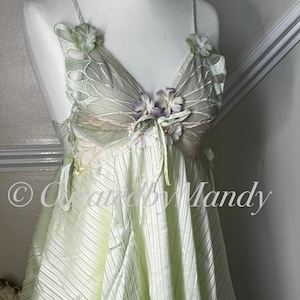 May include: A light green camisole featuring thin spaghetti straps, a V-neckline with sheer butterfly-wing-shaped panels, and decorative fabric flowers in white and purple. The bodice has a pleated texture and a handkerchief hemline.