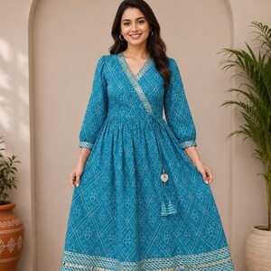 May include: A teal wrap dress featuring a white geometric pattern, three-quarter length sleeves, and a V-neckline. The dress includes a side tie with tassels and a wide metallic gold border along the hem and neckline, reaching mid-calf length.