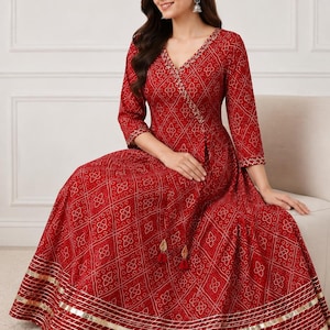 May include: A red floor-length dress featuring a white geometric pattern, a V-neckline, and three-quarter length sleeves. The dress includes a wrap-style bodice with a tie detail featuring red tassels and gold metallic trim along the hemline.