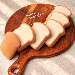 May include: A set of five bread-shaped magnets with light tan centers and darker brown crust edges. The magnets are arranged in a row, and the text Magnetic Snap! is printed above them with a small red horseshoe magnet icon.