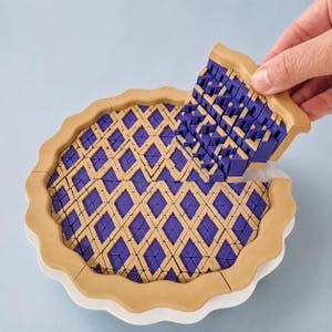 May include: A circular pie-shaped object featuring a tan scalloped crust border and a geometric lattice top pattern. The lattice consists of interlocking tan and purple diamond-shaped segments. One section of the lattice is being lifted upward.