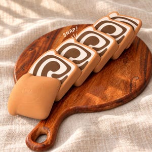 May include: A series of five tan and brown square-shaped silicone cases with a swirl pattern resembling cinnamon rolls, arranged in a row. The word SNAP! is printed in white near the top. Each case features a smooth, matte finish.