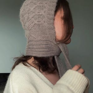 Könnte beinhalten: Eine helltaupefarbene Strickhaube mit einem strukturierten Zopfmuster am Oberkopf und einem gerippten Rand entlang der Gesichtsöffnung. Die Kopfbedeckung verfügt über lange, integrierte Strickbänder, die am Kinnbereich zum Binden verlaufen.