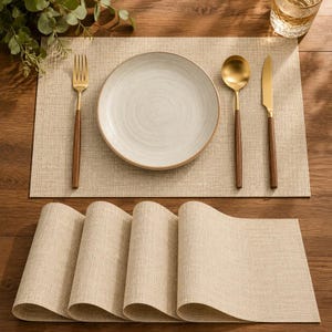 Puede incluir: Un juego de manteles individuales rectangulares de color beige con textura tejida. Un mantel está extendido, con un plato redondo de cerámica color hueso, un tenedor dorado, una cuchara dorada y un cuchillo dorado, cada uno con mango marrón.