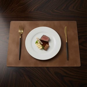 May include: A rectangular brown leather placemat featuring a white ceramic plate centered with a portion of cooked meat, a dollop of mashed potatoes, and a small garnish. A gold and black fork sits to the left, and a gold and black knife to the right.