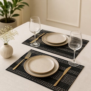 Pode incluir: Um serviço de mesa com dois individuais pretos entrançados, cada um com um prato de jantar e um prato de salada bege salpicado, um garfo dourado, uma faca dourada e um copo de vinho de vidro transparente com relevo estriado.
