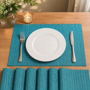 Puede incluir: Un mantel individual rectangular de color verde azulado con una textura fina de rayas verticales. Centrado en el mantel hay un plato llano redondo de color blanco liso. Un tenedor de metal plateado está a la izquierda y un cuchillo a la derecha.