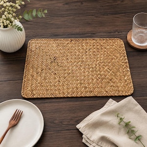May include: A rectangular placemat featuring a woven pattern made from natural light brown dried plant fibers. The mat has rounded corners and a consistent, tight herringbone weave texture throughout its surface, providing a flat base.