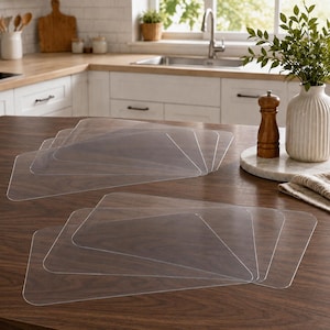 May include: A set of clear, rectangular plastic placemats with rounded corners, arranged in two overlapping stacks on a dark wooden surface. The placemats are transparent, showing the wood grain underneath, and have a smooth, flat finish.
