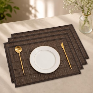 May include: A set of rectangular dark brown bamboo placemats with a woven texture and stitched edges. A white ceramic plate with a thin gold rim is centered on the top mat, accompanied by a gold-toned spoon on the left and a gold-toned knife on the right.