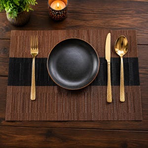Puede incluir: Un mantel individual rectangular tejido en marrón y negro con un plato negro mate centrado, un tenedor dorado con mango martillado, un cuchillo dorado con mango martillado y una cuchara dorada con mango martillado.