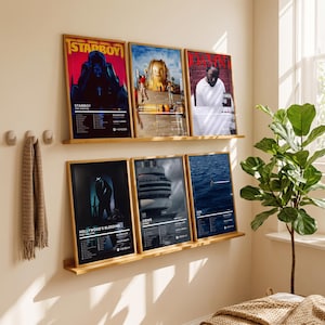 Op de afbeelding: Zes rechthoekige ingelijste muziekalbum-posters op twee houten wandplanken. De posters tonen artwork en tracklists voor Starboy, Astroworld, Damn, Hollywood's Bleeding, Views en SOS, weergegeven in lichte houten lijsten tegen een muur.