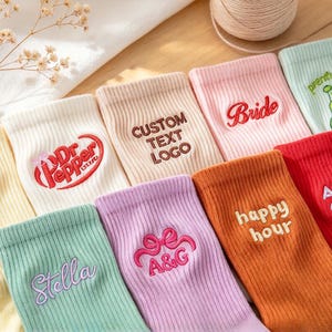 May include: A collection of ribbed crew socks in various colors including yellow, white, beige, pink, light green, lavender, orange, and red. Each sock features unique embroidered text or graphics, including names, logos, and phrases like Bride, Aunt Club, and happy hour.