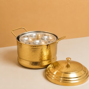 Puede incluir: Una vaporera para idli de metal martillado en color dorado con dos asas laterales y una tapa a juego. El interior contiene una bandeja de metal plateado con cuatro hendiduras circulares para cocinar, con un pomo central.
