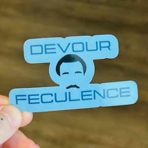 以下が含まれることがあります： 「DEVOUR」と「FECULENCE」の文字がサンセリフ体で書かれた水色のステッカー。口ひげと後退したヘアラインを持つ黒い漫画の顔が特徴です。