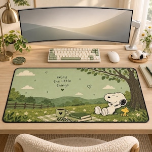 May include: A rectangular desk mat featuring a green landscape illustration with Snoopy and Woodstock relaxing under a tree. The mat includes a picnic blanket, a coffee mug, books, and the text "enjoy the little things" printed in black.