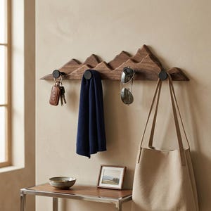 May include: A wall-mounted wooden coat rack shaped like a mountain range with four black circular hooks. The rack holds a set of keys, a folded navy blue scarf, a pair of sunglasses, and a beige canvas tote bag hanging from the hooks.