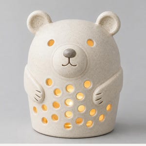Può includere: Una lampada in ceramica color beige maculato a forma di orso, con fori circolari ritagliati sul corpo e sul viso. L'orso presenta piccole orecchie, un muso arrotondato con un naso scuro e braccia che riposano lungo i fianchi.