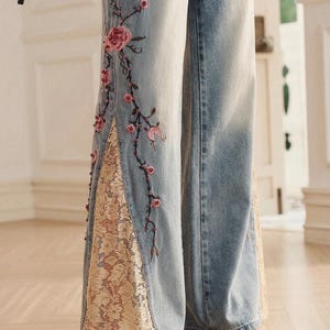 May include: Light blue denim jeans featuring wide legs with side slits. The jeans are decorated with pink floral embroidery and bead accents along the side seams, complemented by beige lace inserts within the slits. The logo TH THE HAILLEY JEANS is visible.