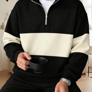 Könnte beinhalten: Ein schwarzer Langarm-Pullover mit Viertelreißverschluss und einem breiten, cremefarbenen horizontalen Streifen über Brust und Ärmel. Das Kleidungsstück hat einen hohen Kragen, gerippte Bündchen und ein weißes Unterhemd am Halsausschnitt.