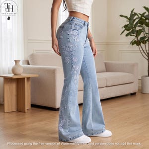 May include: Light blue high-waisted flare jeans featuring white floral embroidery along the side of the leg. The denim trousers have a classic five-pocket design, a button and zip closure, and are paired with white trainers and a white crop top.