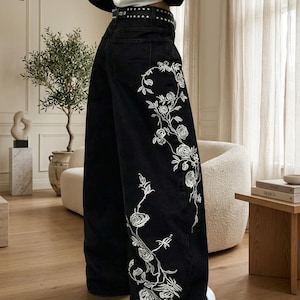 Op de afbeelding: Zwarte jeans met wijde pijpen en witte bloemenborduursels die vanaf de zoom langs de zijkant omhoog lopen. De broek heeft een hoge taille met een zwarte riem met studs en wordt gecombineerd met een wit en zwart shirt met lange mouwen.