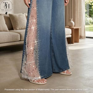 Peut inclure: Jean large en denim bleu doté de panneaux latéraux verticaux transparents en maille blanche. Les panneaux en maille sont ornés de paillettes argentées éparpillées et de petites perles blanches, créant un effet scintillant le long des jambes.