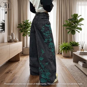 May include: High-waisted black denim jeans featuring a wide-leg silhouette and a black belt with silver studs. The side of the jeans is decorated with an intricate, embroidered pattern of teal-colored roses and winding vines extending downward.