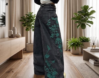 May include: High-waisted black denim jeans featuring a wide-leg silhouette and a black belt with silver studs. The side of the jeans is decorated with an intricate, embroidered pattern of teal-colored roses and winding vines extending downward.