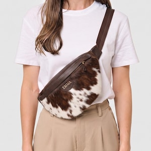 May include: A brown and white cowhide crossbody bag featuring a dark brown leather top panel with a gold zip closure. The front of the bag displays the name EMMA in gold lettering. It includes an adjustable dark brown fabric strap.