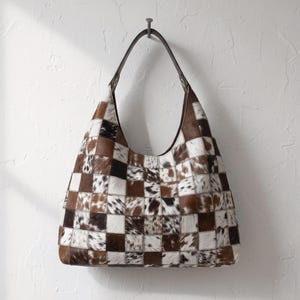 Könnte beinhalten: Eine Hobo-Handtasche mit einem Patchwork-Design aus quadratischen Feldern in verschiedenen Braun-, Weiß- und Schwarztönen aus Kuhfell. Die Tasche hat einen einzelnen dunkelbraunen Leder-Schulterriemen mit Metallbeschlägen.