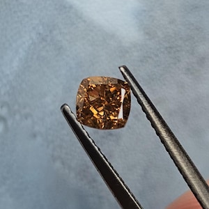 Può includere: Una pietra preziosa taglio cuscino di colore marrone cognac intenso con superficie sfaccettata, tenuta da una pinzetta in metallo scuro. La pietra mostra riflessi di luce interni e bordi netti, esibendo un aspetto chiaro, trasparente e lucido.