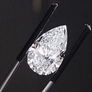 May include: A pear-shaped, colourless, faceted gemstone held by a pair of metal tweezers with black rubber-coated tips. The stone exhibits high clarity and brilliance, reflecting light through its numerous triangular and kite-shaped facets.