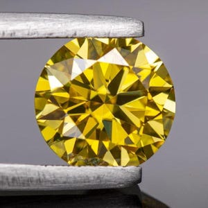 Op de afbeelding: Een ronde, briljant geslepen gele edelsteen vastgehouden door een zilverkleurige metalen pincet. De steen heeft een complex geometrisch facetpatroon dat licht reflecteert, wat zorgt voor een levendige, verzadigde gele kleur over het hele oppervlak.