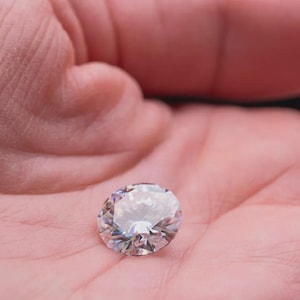 Könnte beinhalten: Ein runder, im Brillantschliff gehaltener, klarer Edelstein mit zahlreichen Facetten, die das Licht reflektieren und für ein funkelndes Aussehen sorgen. Der Stein ist farblos, transparent und weist ein geometrisches Muster aus dreieckigen Facetten auf.