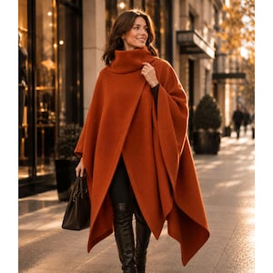 May include: A burnt orange poncho featuring a high cowl neckline and an asymmetrical draped front design. The garment has a smooth, solid texture and falls in loose, flowing panels. It is paired with black leggings, black boots, and a black handbag.
