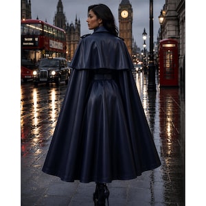 May include: A floor-length, dark navy blue coat with a cape-style overlay and a belted waist. The garment features a smooth, structured finish and a flared silhouette that extends to the ankles, worn over dark footwear in a city street setting.