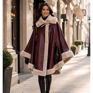 May include: A deep burgundy leather cape featuring a wide, cream-colored faux fur collar and matching faux fur trim along the front opening and hem. The garment includes a toggle closure at the neck and is worn with black leather gloves and boots.