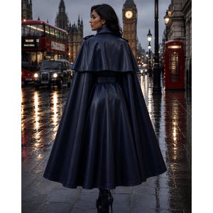 May include: A floor-length, dark navy blue coat with a cape-style overlay and a belted waist. The garment features a smooth, structured finish and a flared silhouette that extends to the ankles, worn over dark footwear in a city street setting.