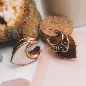 May include: A pair of ear gauges featuring a crescent moon shape with a metallic finish. One gauge has a smooth, iridescent white inlay, while the other displays a detailed, sunburst-style engraved pattern on the metallic surface.