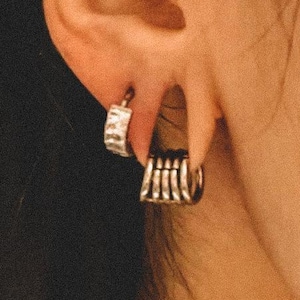 May include: Two silver-toned hoop earrings worn in the earlobe. The upper earring is a small, textured hoop, while the lower earring features a multi-layered, ribbed design consisting of several thin, connected silver-toned bands.