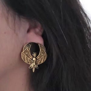 May include: A circular ear plug featuring a black center and a gold-toned metal overlay. The overlay depicts a detailed moth with spread wings, a skull at the center of its body, and intricate celestial patterns including crescent moons on the wings.